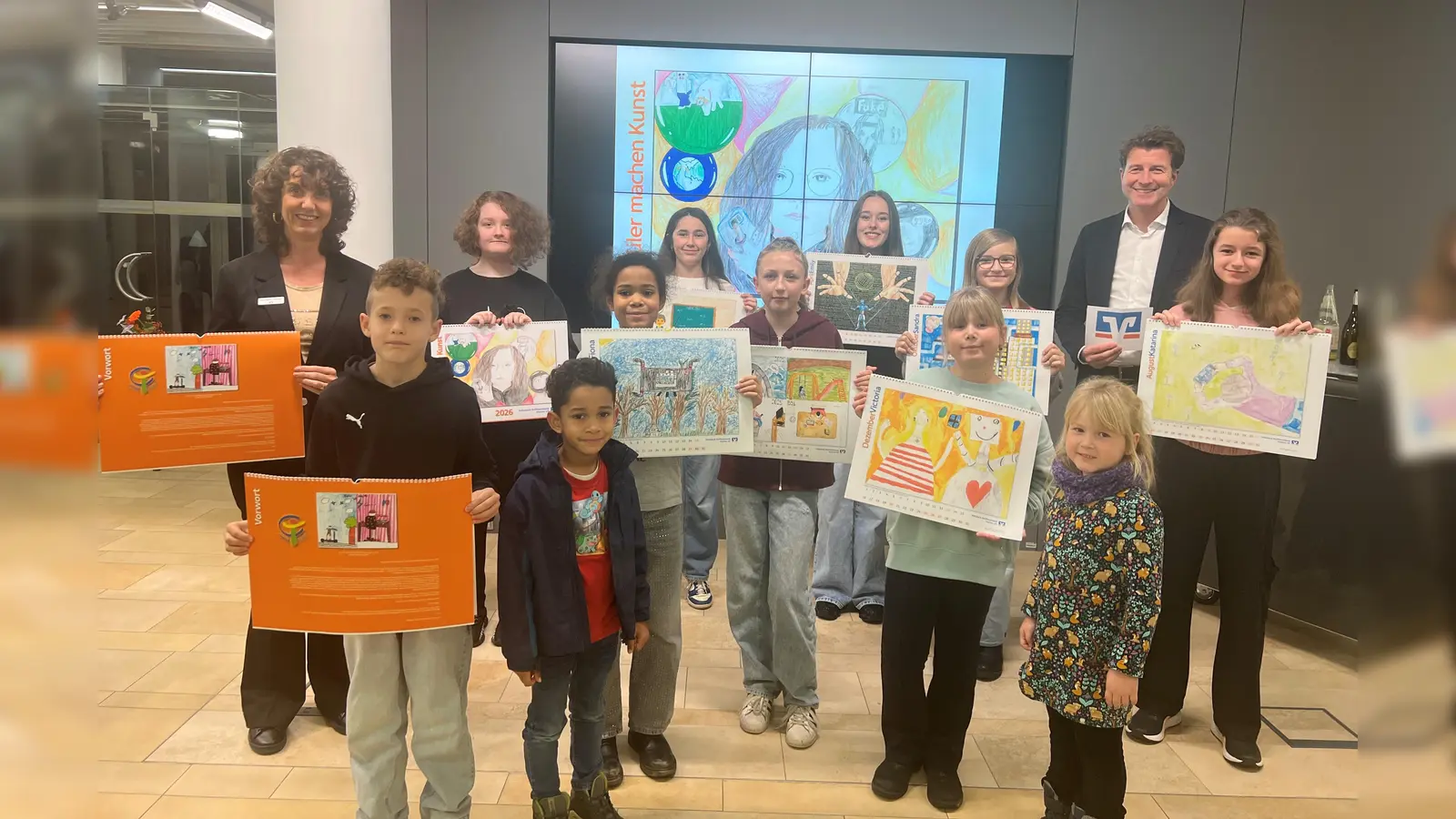 Stolz präsentieren die Kinder und Jugendlichen den Kalender 2026 „Schüler machen Kunst“ zusammen mit Bankvorstand Klaus Berger (r.) und Marketingreferentin Michaela Steiner (l.). (Foto: VR Bank Dachau)